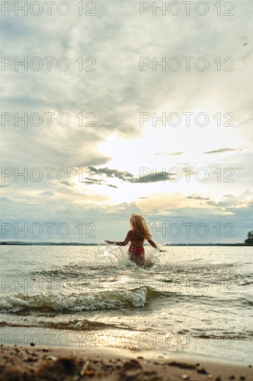 Funny woman plays joyfully in the water at a lakeside beach. The sun sets behind her, casting