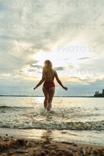 A woman in a red swimsuit steps into a tranquil lake, creating splashes as the sun sets behind her.