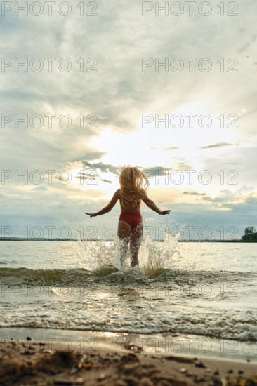 A woman in red swimwear plays in the water, splashing joyfully as the sun sets in the background.