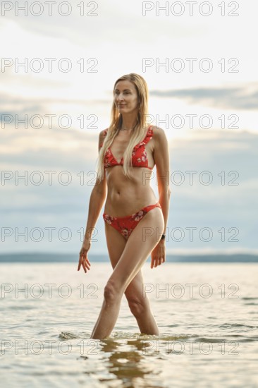 A woman strolls gracefully through shallow waters at sunset, wearing a vibrant red bikini adorned
