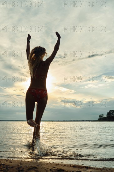 A woman runs through shallow water, joyfully throwing up her arms towards the setting sun