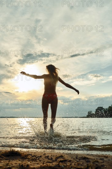 A woman wearing swimwear joyfully splashes through water at sunset. The sky displays vibrant