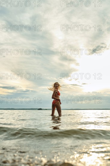 A woman wades through shallow water, smiling as she enjoys the warm sunset. The sky is painted with