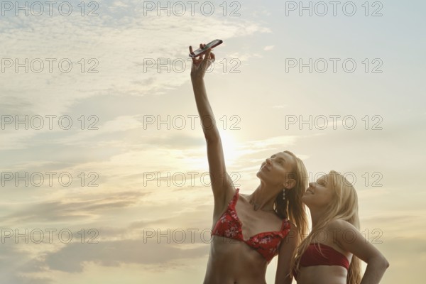 Two friends in swimwear stand on the beach, smiling as they take selfies. The sun sets behind them,