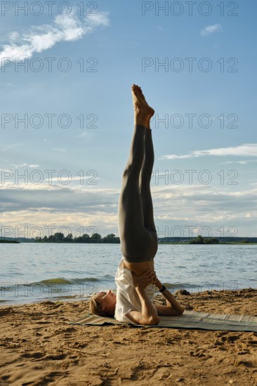 A woman performs shoulder blade stand with her legs raised high at a lakeside on a sandy shore. The