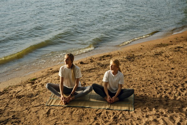 Two women are sitting on a mat by the water's edge and doing yoga poses as the sun begins to set.