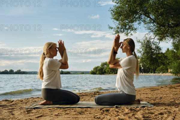 Two women sit on a mat in front of each other by the water, practicing yoga. They perform a