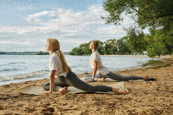Two women engage in yoga poses on the sandy beach beside a calm lake. The sun shines brightly,