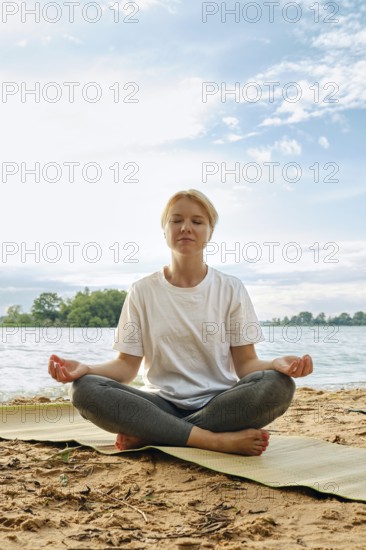 A woman sits cross-legged on a mat by the lake, eyes closed in meditation. The sun shines down on