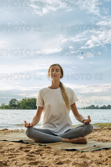 A woman sits cross-legged on a mat by the lake, practicing yoga in a serene environment. The sun