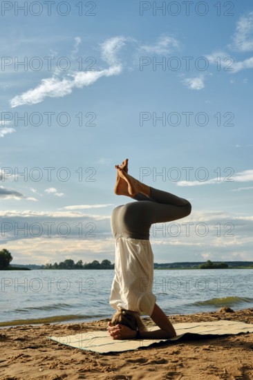 A slender woman performs a headstand yoga pose on a mat in the sand near a tranquil lake. The