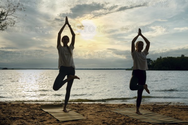 Silhouettes of two women performing yoga poses on mats along a sandy lakeshore as the sun sets,