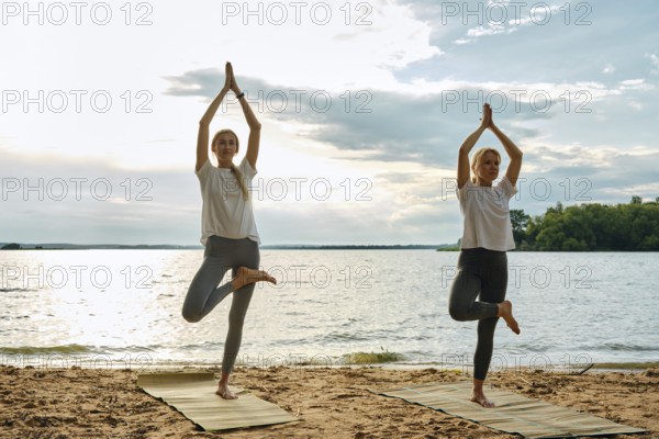 Two women perform yoga poses on mats at the beach during sunset. They are surrounded by a tranquil