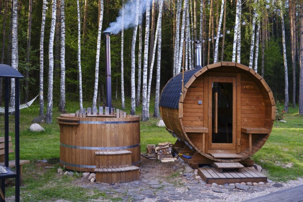 A wooden sauna and a hot tub are nestled in a lush forest. The hot tube emits smoke from its