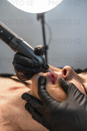 Beautician wearing black gloves applying permanent makeup on lips using a tattoo machine in a