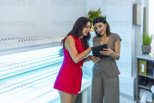 Two beauticians are consulting a digital tablet, discussing treatment options near a solarium,