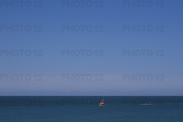 Sailboat, colourful sail, sailing on sea, sky, blue, behind offshore wind farm, wind turbines,