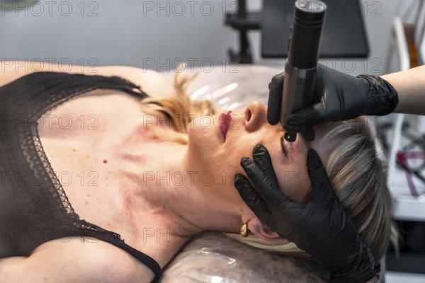 Beautician applying permanent eyeliner makeup on upper eyelid of client lying on bed in beauty