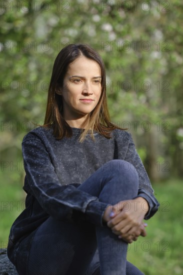 A woman sits quietly in an outdoor area, surrounded by lush greenery and soft light. She appears