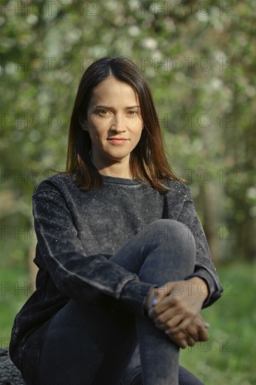 A young woman with long dark hair sits comfortably on a rock, wearing a casual sweater and leggings