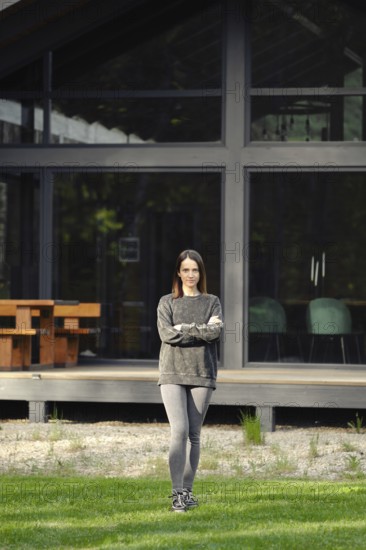 A young woman stands crossing her hands on the lawn in front of her modern house with large glass