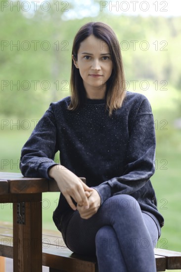 A woman is seated on a wooden bench, dressed in a black sweatshirt and leggings. She gazes directly