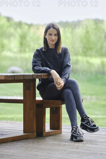 A young woman sits on a wooden bench beside a table, wearing casual attire. She enjoys the peaceful