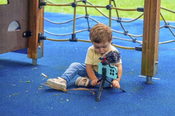 Little child sitting on playground's rubberized floor setting up a smartphone on a tripod with a