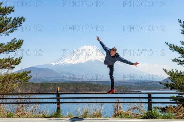 Young man balancing on railings next to a road, view across the lake to Mt Fuji volcano, Motosu