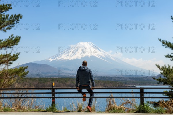 Young man sitting on railings next to a road and looking across the lake to Mt Fuji volcano, Motosu