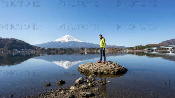 Young man standing on a rock in water, volcano Mt. Fuji is reflected in Lake Kawaguchi, Yamanashi