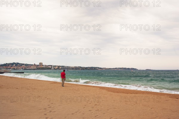 Man wearing a red shirt looking at the moving sea, Fréjus in the background, Provence-Alpes-Côte
