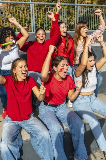 Young women sitting on stadium bleachers, cheering loudly with raised arms and painted faces,