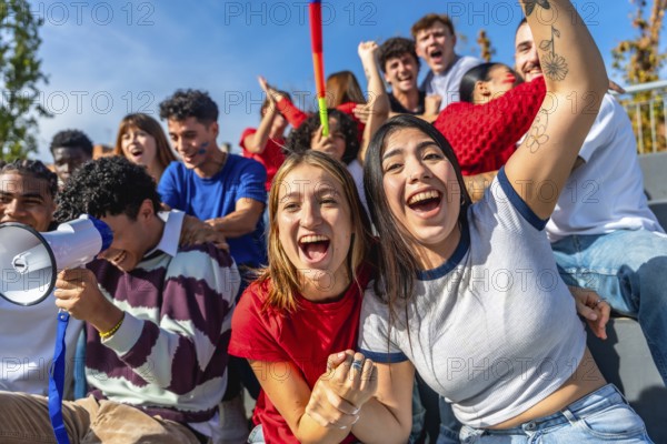 Group of excited young people watching a sports game and cheering with hands up and megaphones,