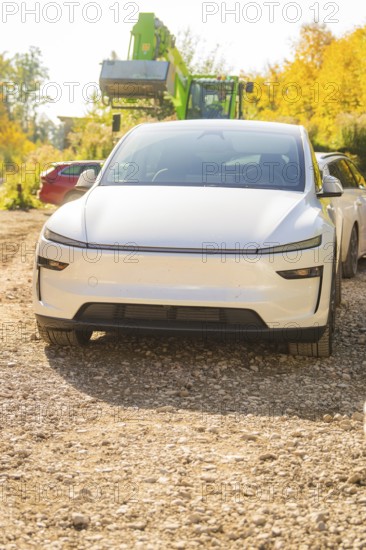 White electric car on a gravel path in autumn, surrounded by colorful foliage and trees, Telsa