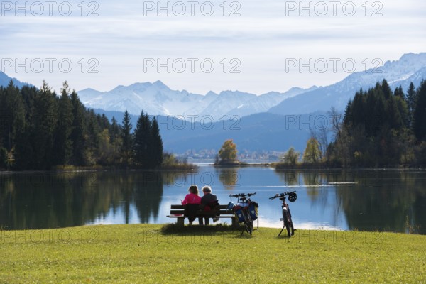 Two woman sitting on a bench look at the peaceful Forggensee with mountain views, surrounded by