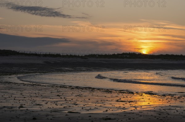 Sunset on a quiet beach with soft light on the waves, wet beach at low tide, dunes in the