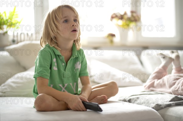 Child, boy, 7 years old, blonde, sitting on sofa, with remote control in hand, watching TV,