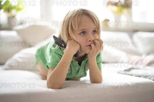Child, boy, 7 years old, blonde, lying on sofa with remote control in hand, watching TV, watching