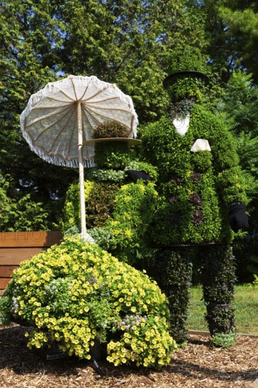 Live plant sculpture of Victorian man standing and woman with sun shade umbrella sitting on wooden