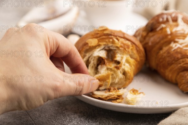 Croissant on white plate with hand on brown concrete background and linen textile, cup of coffee,