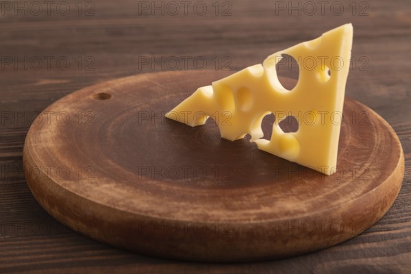 Piece of cheese with holes on cutting board on brown wooden background, side view, close up,