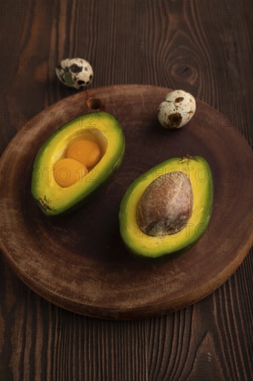 Two Avocado halves with pit and Egg yolk on round wooden cutting board on brown wooden, copy space,