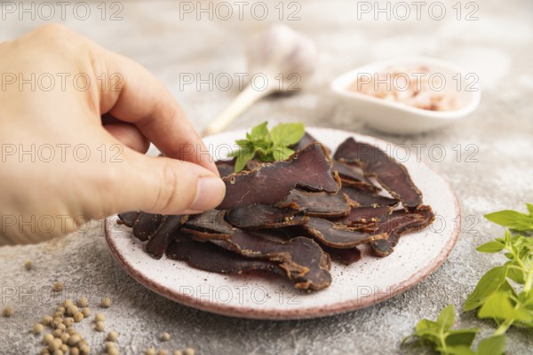 Armenian Basturma dried meat with hand on plate with pepper and herbs on brown concrete background.