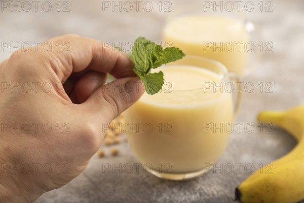 Organic non dairy banana and soy milk in glass with hand on brown concrete background. Vegan