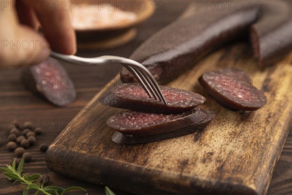 Sujuk sausage with hand on wooden cutting board with pepper and herbs on brown wooden background.