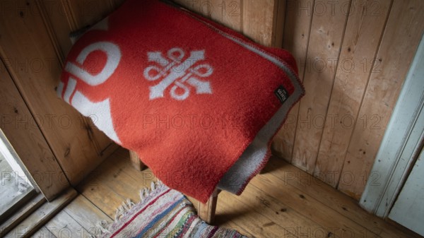 Rote Wolldecke mit Olavsweg-Symbol liegt auf einem Holzschemel, historischer Bauernhof und