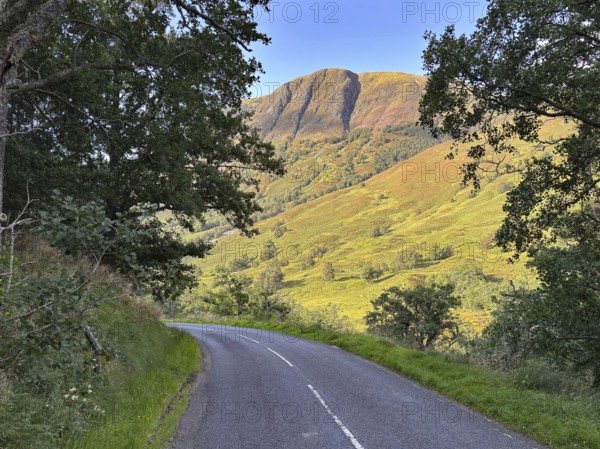 A winding road leads through a green landscape under a clear sky with a mountain in the background,