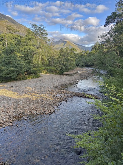 A tranquil river flows through a wooded landscape with mountains in the background and a clear blue