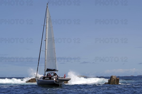 A sailing boat navigates through rocky sea with blue sky in the background, Isles of Scilly,